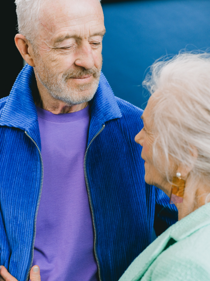 Older adult male wearing a purple t-shirt and blue jacket looking lovingly at older adult female wearing a light green jacket