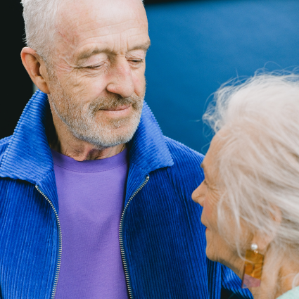 Older adult male wearing a purple t-shirt and blue jacket looking lovingly at older adult female wearing a light green jacket