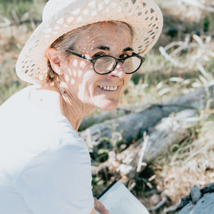 Older adult woman wearing a straw hat and glasses. She appears to be looking up from a book that she is reading while sitting outdoors.