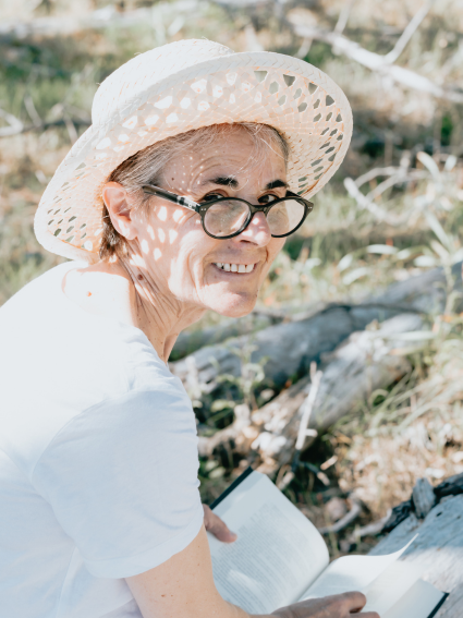 Older adult woman wearing a straw hat and glasses - looking up and smiling as she enjoys a book while sitting outside in nature.