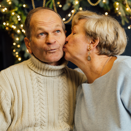 Older adult woman in light blue sweater kissing an older adult male on the cheek. The couple is sitting in front of a lighted Christmas tree