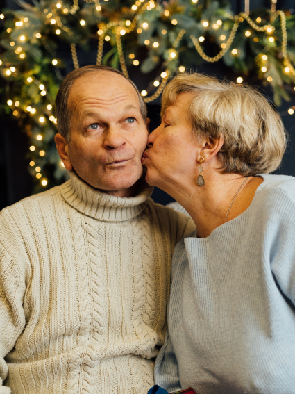 Older adult woman in light blue sweater kissing an older adult male on the cheek. The couple is sitting in front of a lighted Christmas tree